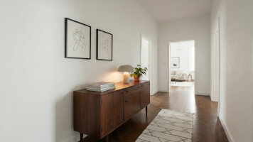 Kategorie Wände und Decken - Heller Flur mit weiß gestrichenen Wänden und glattem Holzboden. Auf einer Kommode stehen eine Tischlampe, ein Stapel Zeitschriften und eine grüne Topfpflanze. An der Wand hängen zwei gerahmte abstrakte Linienzeichnungen.