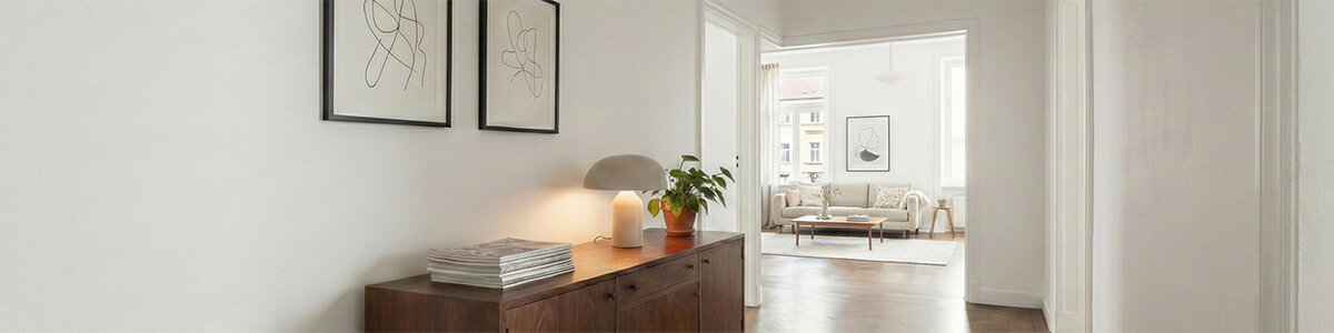 Heller Flur mit weiß gestrichenen Wänden und glattem Holzboden. Auf einer Kommode stehen eine Tischlampe, ein Stapel Zeitschriften und eine grüne Topfpflanze. An der Wand hängen zwei gerahmte abstrakte Linienzeichnungen. Im Hintergrund ist ein Wohnzimmer mit großen Fenstern und einer hellen Sitzgruppe zu sehen.