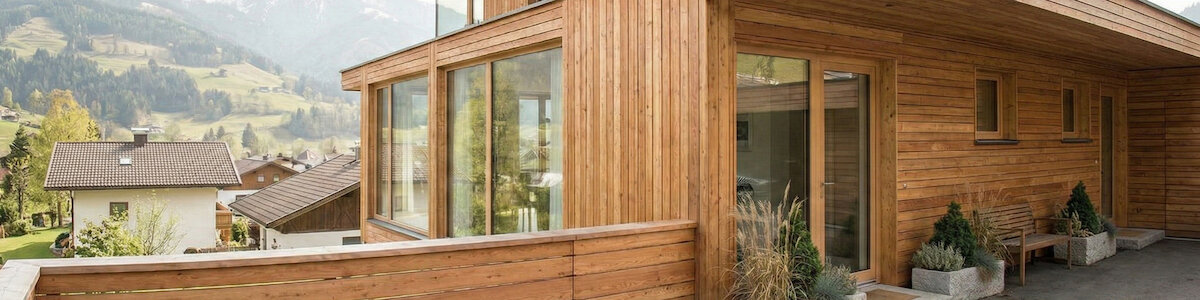 Schöner Eingangsbereich eines Holzhauses. Vor grüner Bergkulisse. An der Wand stehen verschiedene Blumentöpfe genauso wie eine Bank.